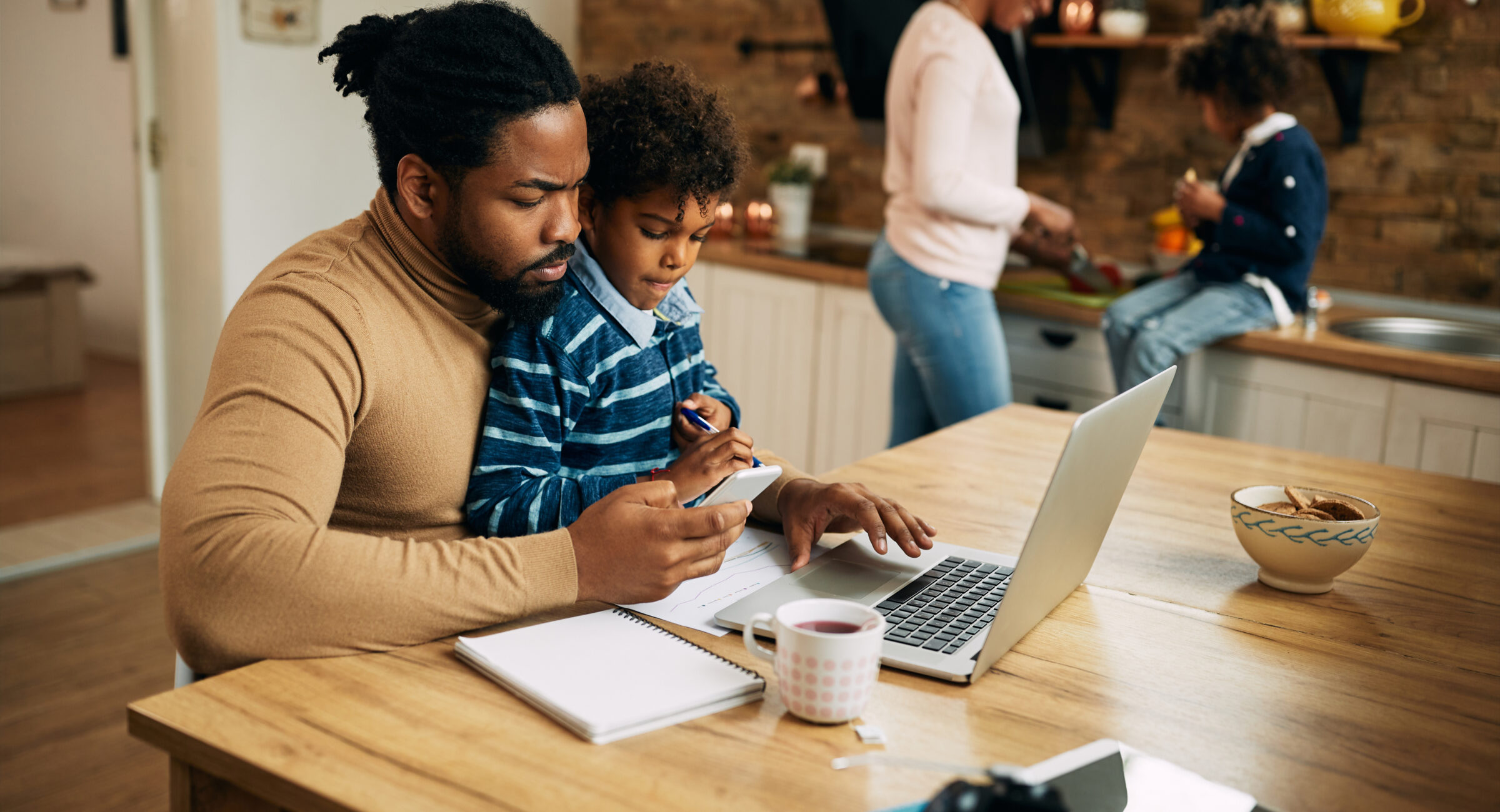 Black working father using laptop and mobile phone while son is sitting on his lap. Mother and daughter are in the background.